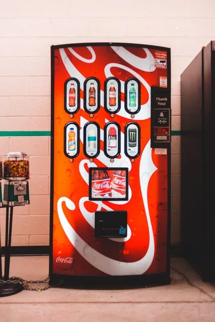 Colorful coin operated soda machine with symbols of assorted bottles and coke near small candy machine on floor near tiled wall