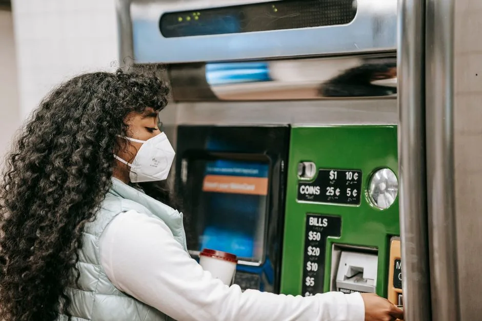 Side view of black lady in casual outfit with protective mask and takeaway cup with drink standing in metro while putting card in ticket machine