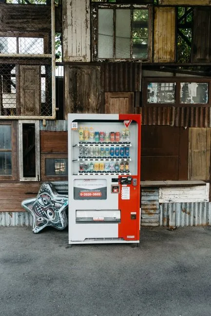 An old vending machine in front of a rustic wall with various drinks on display.