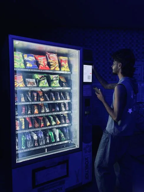 A man selecting snacks from an illuminated vending machine indoors at night.