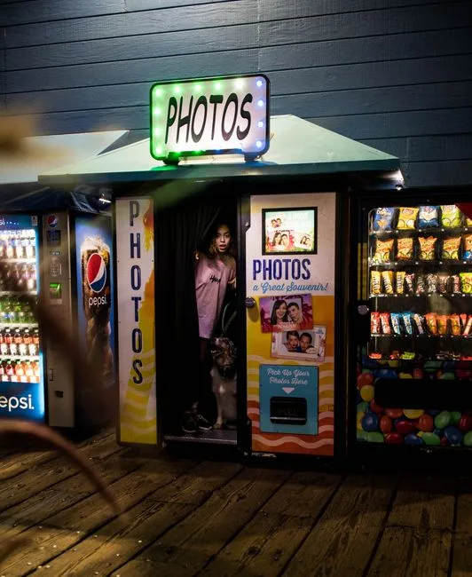 A woman and a dog standing inside a vintage photobooth at night surrounded by vending machines.