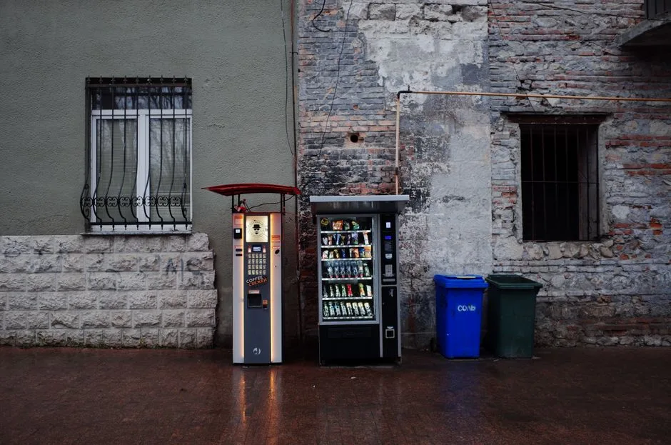 Vending machines and trash bins against an urban stone wall backdrop.