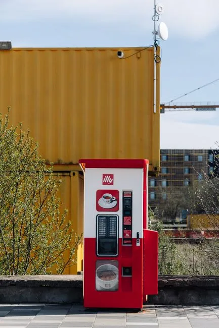 Bright red coffee vending machine next to a yellow container outdoors under a clear blue sky.