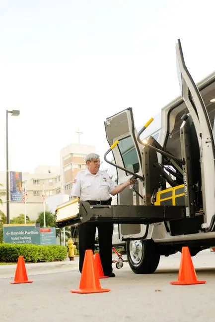 A security guard operates a mechanical lift on a van in a parking area, surrounded by traffic cones.