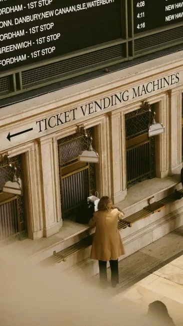 A person in a trench coat stands at a ticket counter in a busy train station.