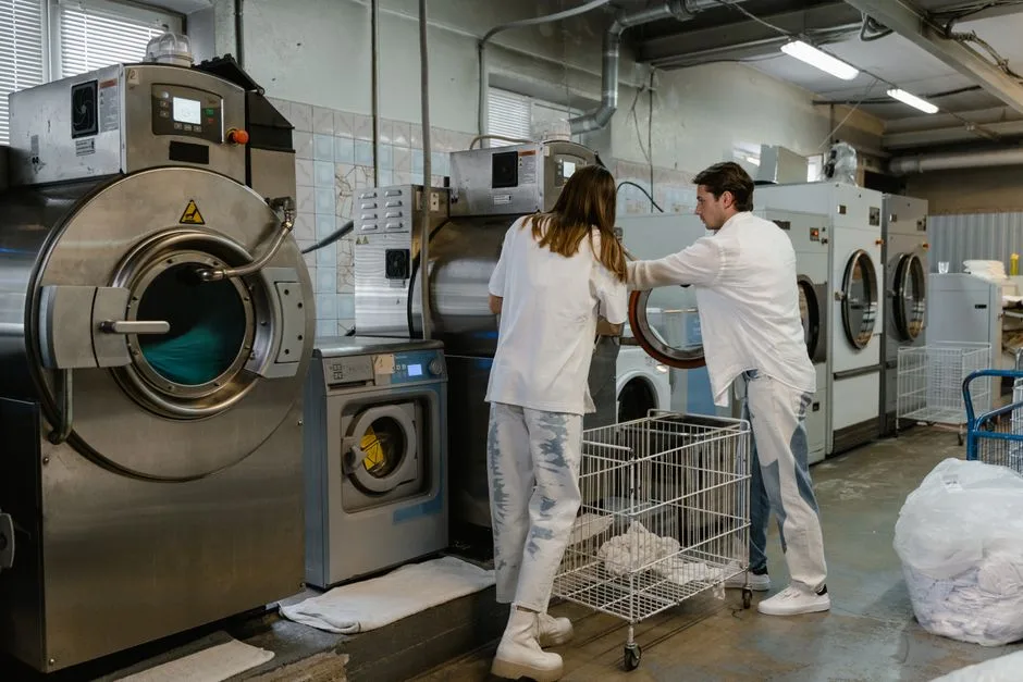 Two adults working in an industrial laundry facility with large machines.